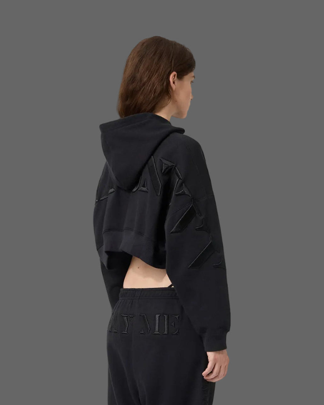 A model shown from the back wearing a black cropped hoodie with geometric panel details on the sleeves and a matching black sweatpants, set against a solid dark gray background.